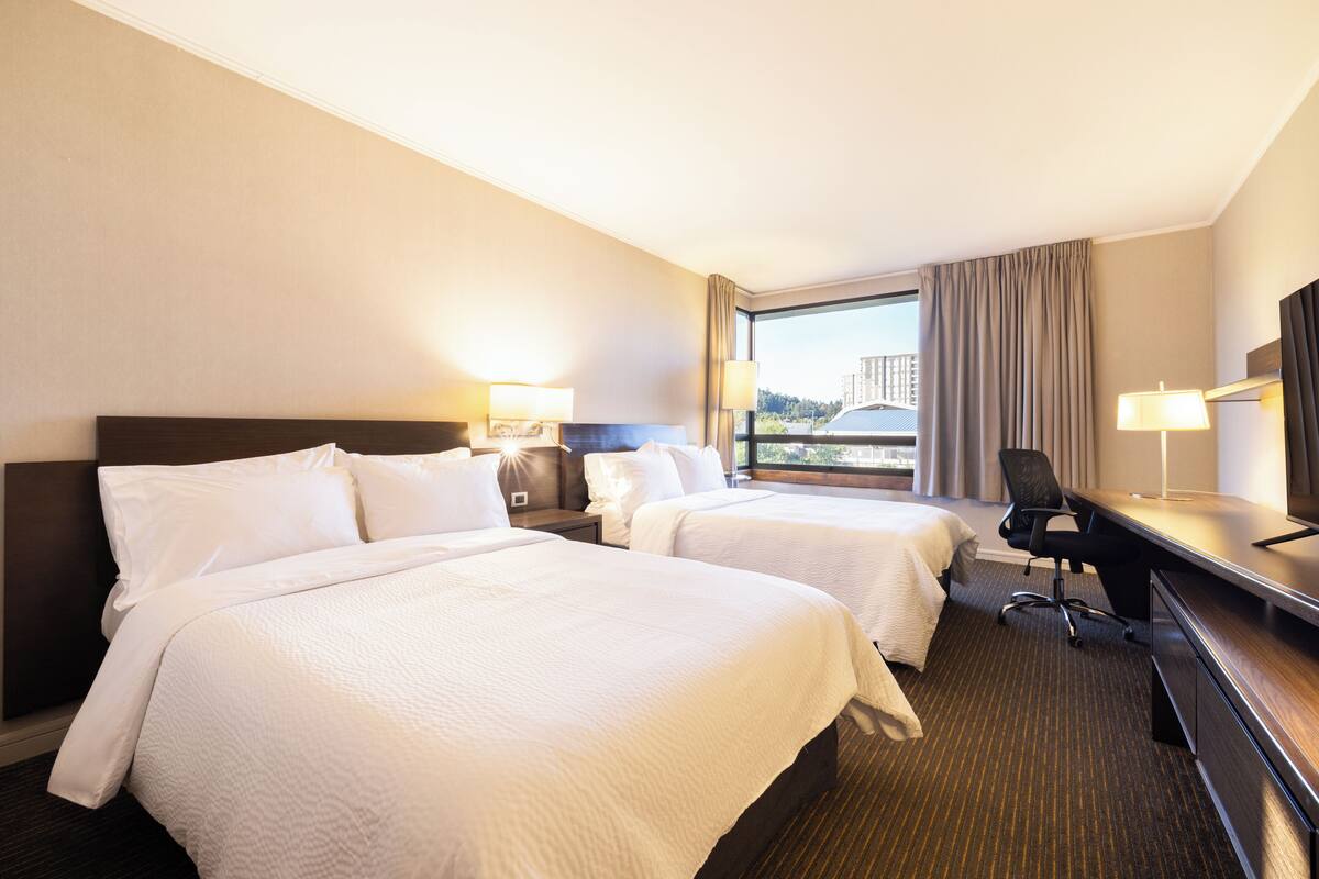 Une chambre d’hôtel avec deux lits, un bureau et une chaise. Il y a une fenêtre avec des rideaux, une lampe et une vue sur un bâtiment à l’extérieur.