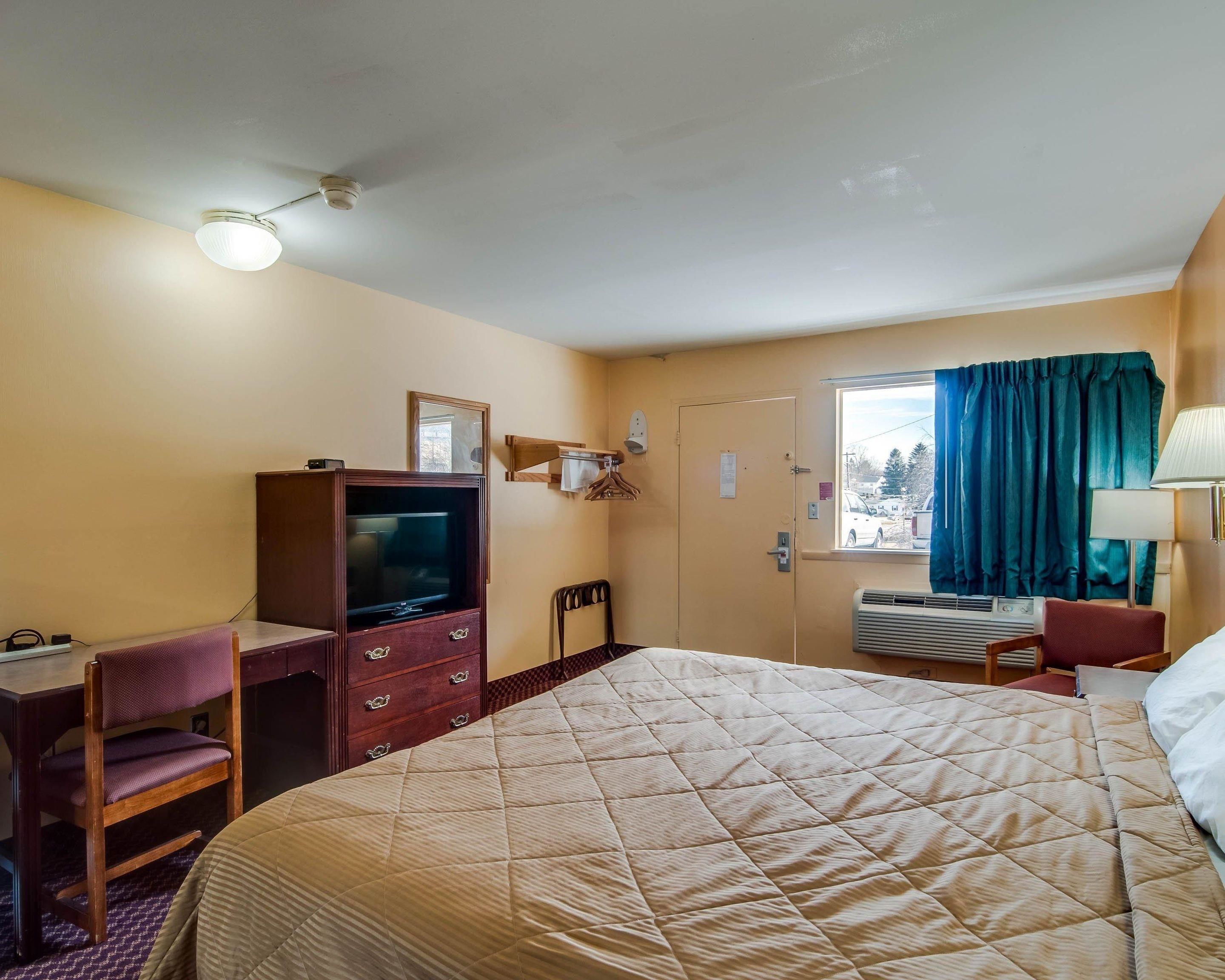 Une chambre d’hôtel comprenant un lit, un bureau avec une télévision, une chaise, une commode, une fenêtre avec des rideaux et un ventilateur de plafond.
