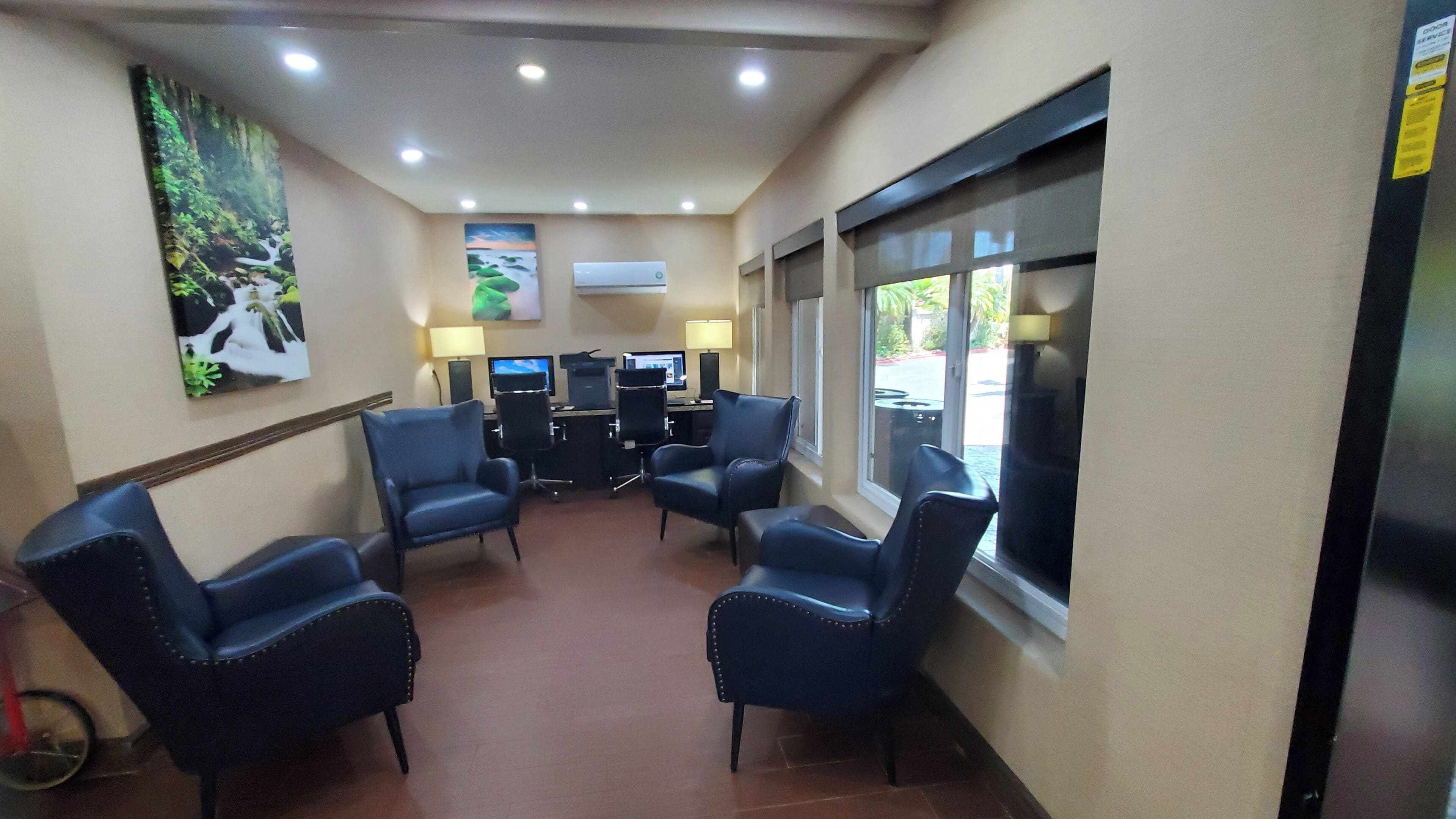 A hotel room with a seating area, two computers, a window, and a painting on the wall.