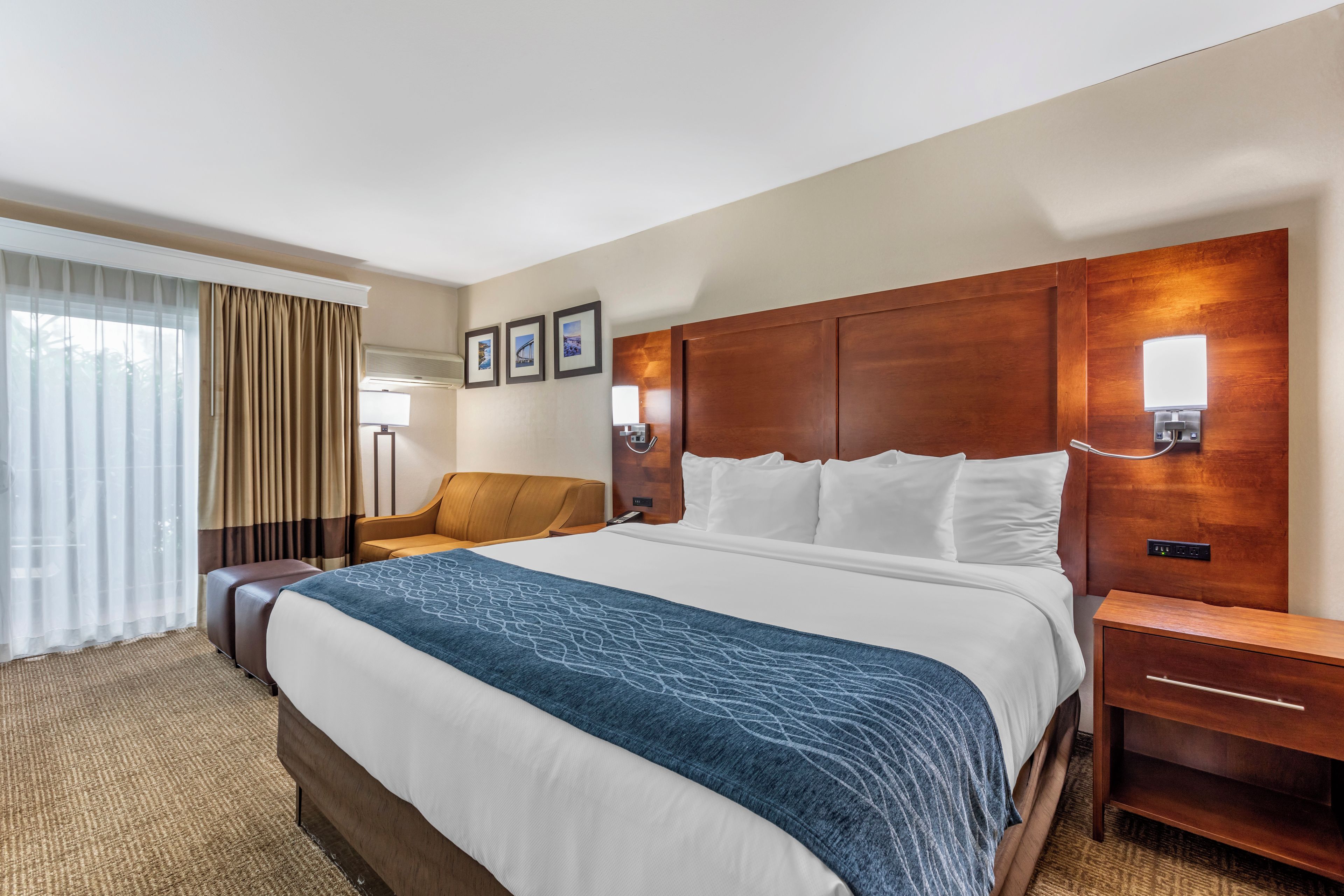 Quarto de hotel com uma cama grande, cabeceira de madeira, mesa de cabeceira, uma cadeira e uma janela com cortinas.