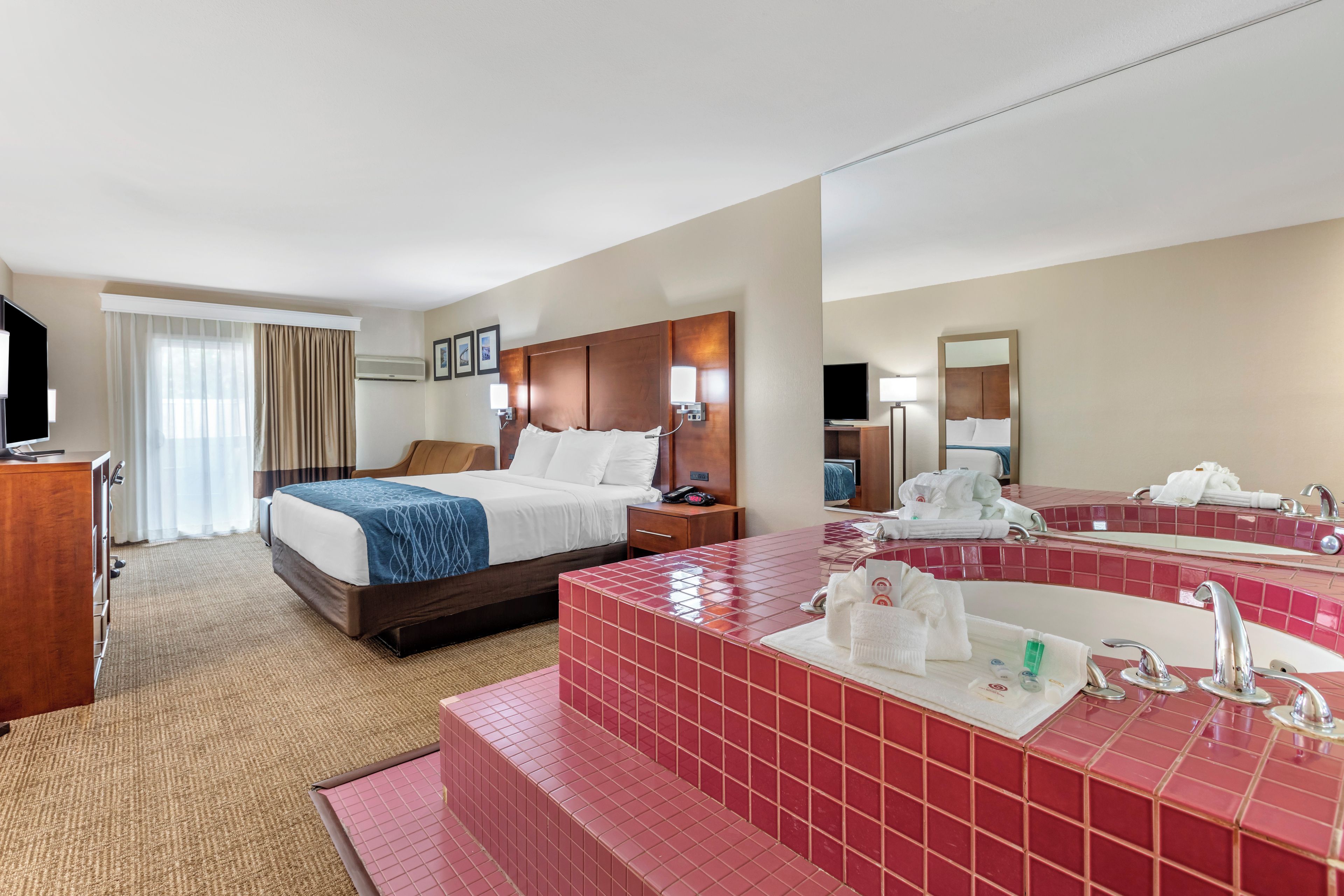A hotel room with a bed, a desk, a television, a bathroom with a large mirror and red-tiled sink.