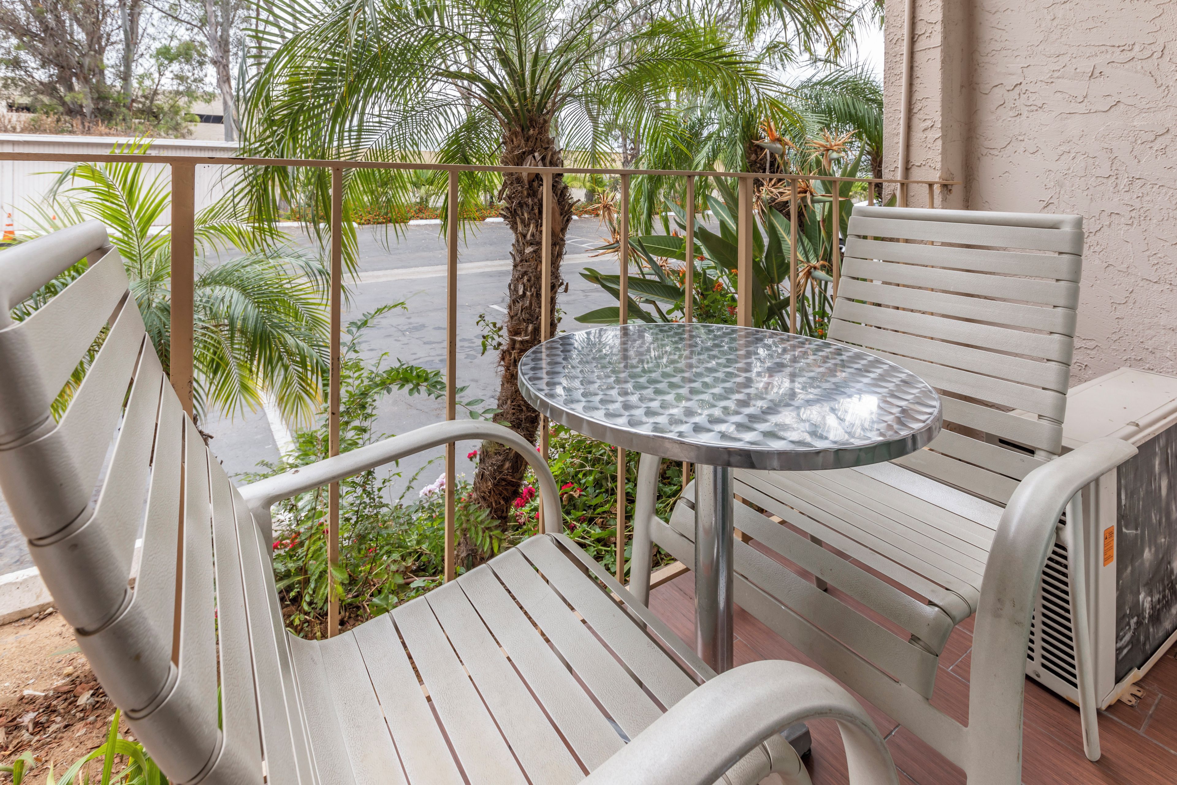 A balcony with a metal table and two white chairs, surrounded by greenery and a palm tree.