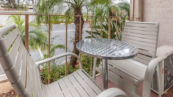 A balcony with a metal table and two white chairs, surrounded by greenery and a palm tree.
