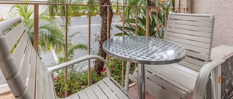 Un balcón con una mesa de metal y dos sillas blancas, rodeado de follaje y una palmera.