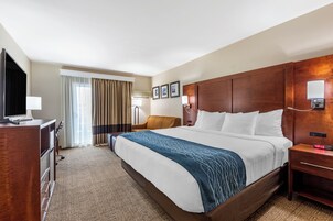 Una habitación de hotel con una cama grande, un escritorio con televisor de pantalla plana, una silla y una mesita de noche con lámpara.