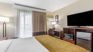 A hotel room with a bed, a television, a desk with a chair, a refrigerator, and a window with curtains.