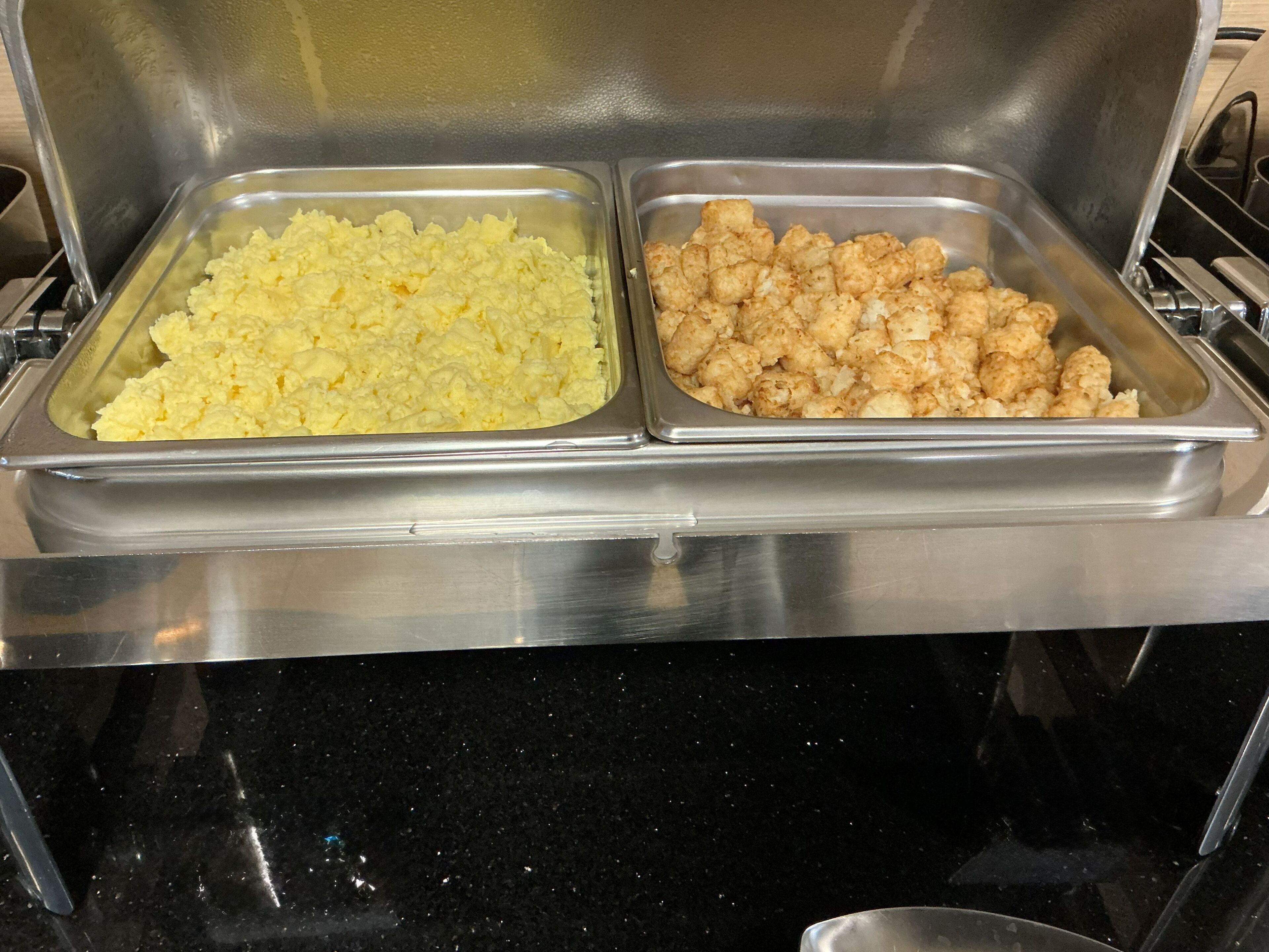 A buffet with two metal trays, one containing scrambled eggs and the other containing fried chicken.
