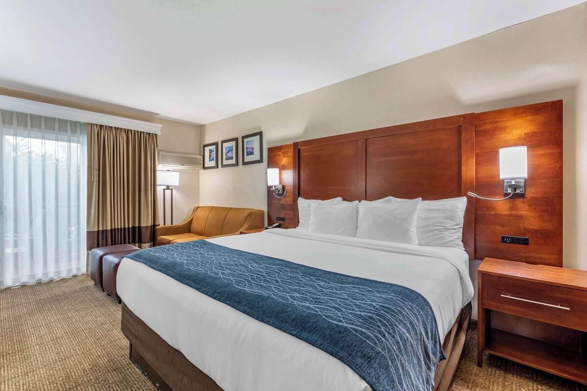 Camera d'albergo con un letto grande, una testiera in legno, un comodino, una scrivania, una sedia, una finestra con tende e luci a parete.