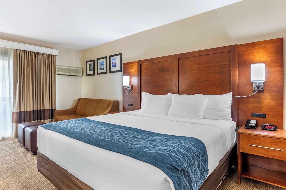 Ein Hotelzimmer mit einem großen Bett, einem hölzernen Kopfteil, einem Nachttisch mit Lampe, einem Sessel und einem Fenster mit Vorhängen.