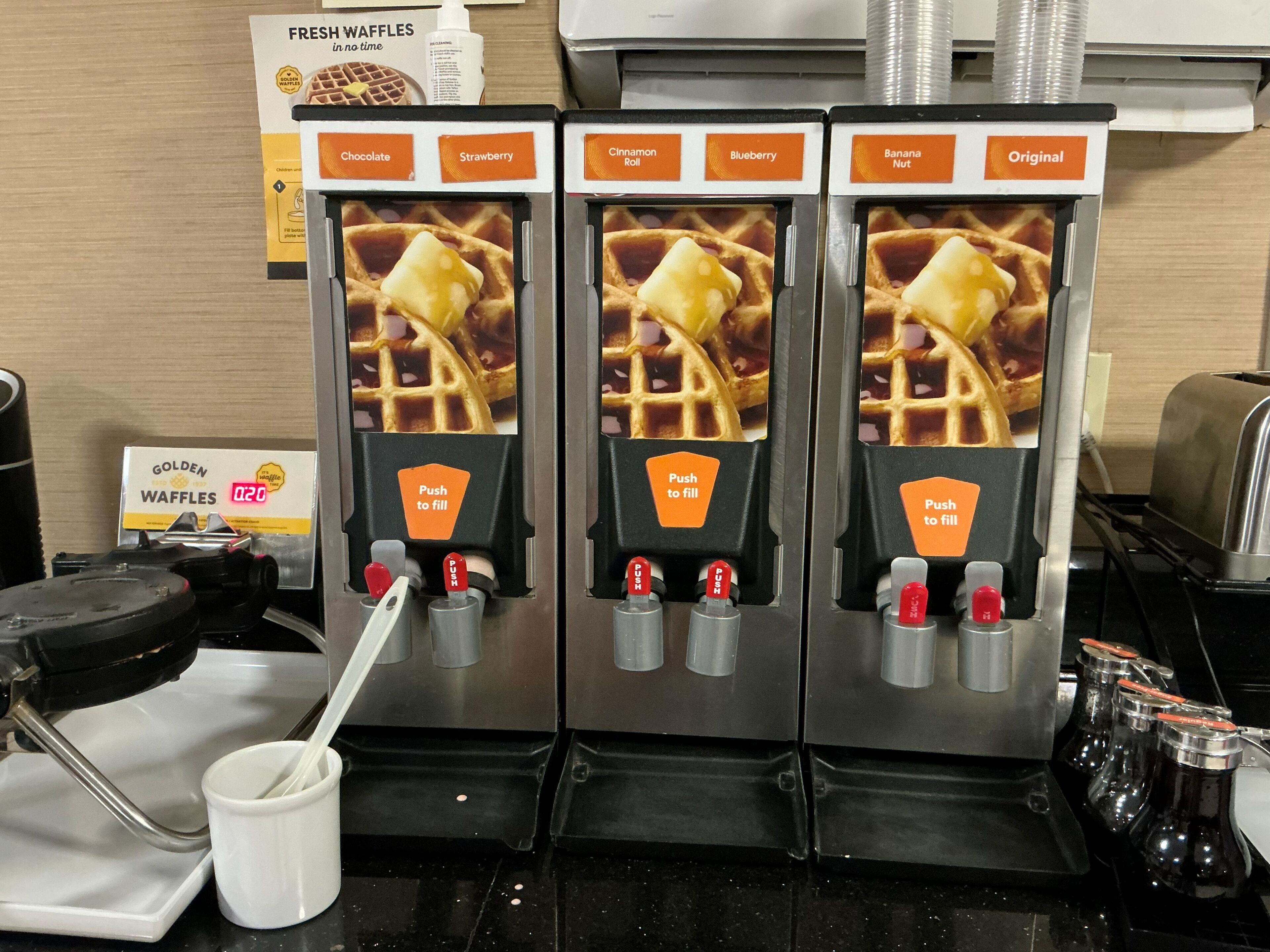 Una macchina per waffle con diverse opzioni di sapore, tra cui cioccolato, fragola, cinnamon roll, mirtillo, banana e noci, e gusto originale.