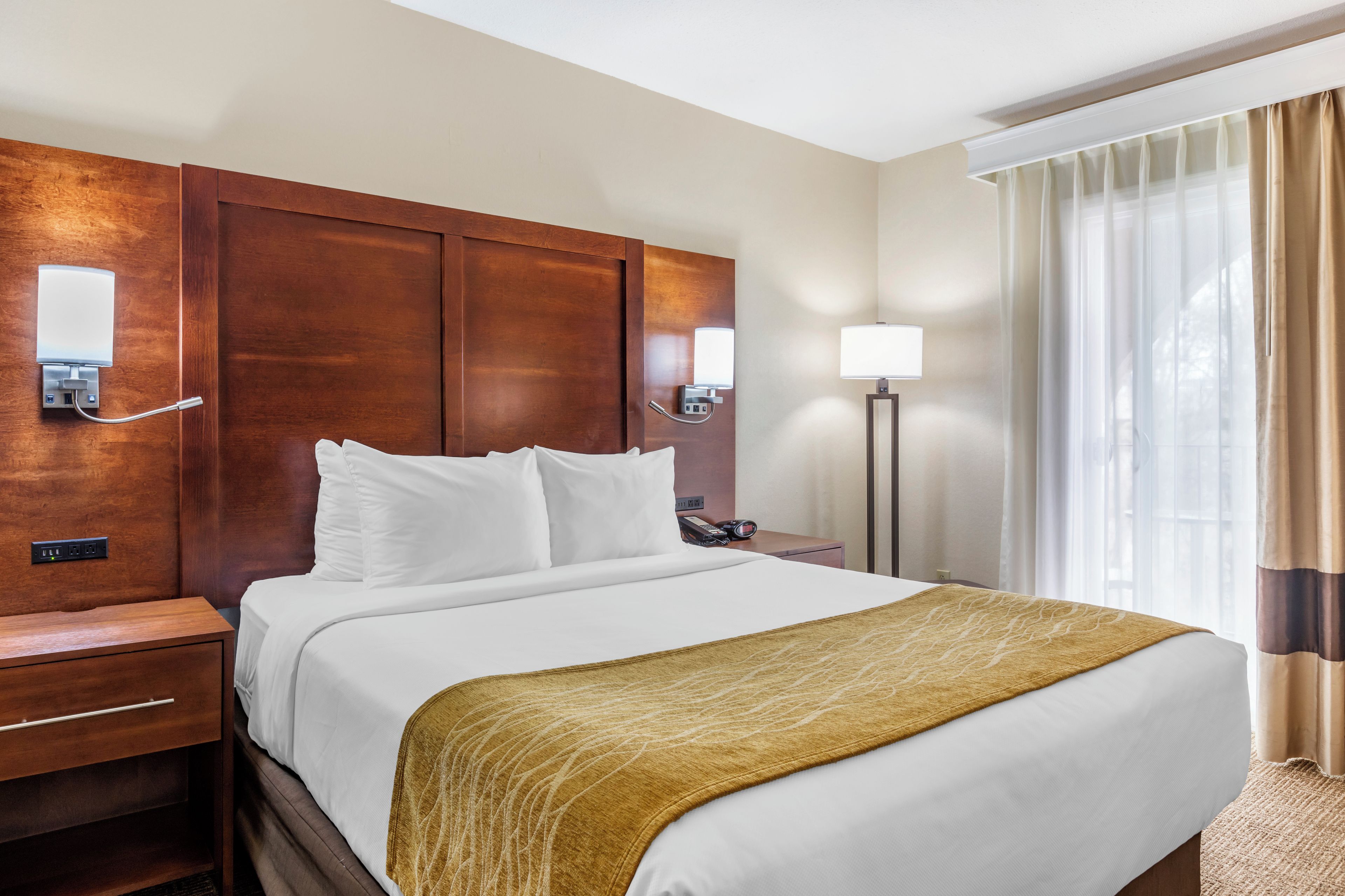 Ein Hotelzimmer mit einem großen Bett, einem Holzkopfteil, einem Nachttisch mit Lampe und einem fenster mit Vorhängen.