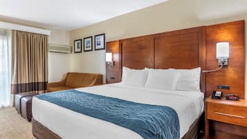 Habitación de hotel con una cama grande, un cabecero de madera, una mesita de noche con lámpara, una silla y un equipo de aire acondicionado.