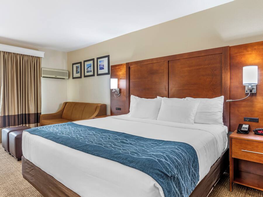 A hotel room with a large bed, a wooden headboard, a bedside table with a lamp, a chair, and an air conditioning unit.