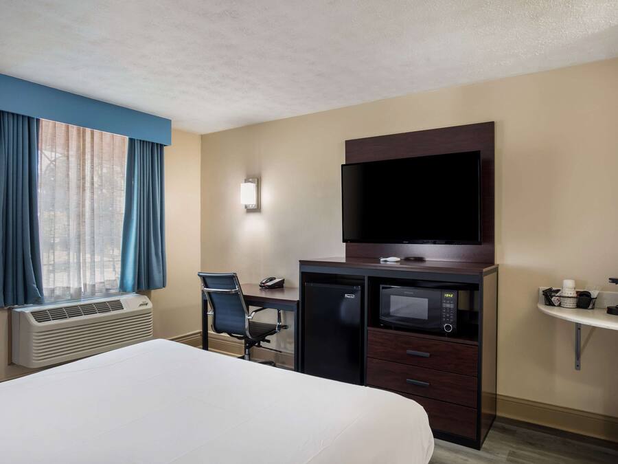Ein Hotelzimmer mit Bett, Schreibtisch, Stuhl, Klimaanlage, Fernseher, Mikrowelle und einem kleinen Regal mit Gegenständen.