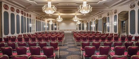 A conference room with rows of red chairs, chandeliers, and ornate wall decorations.