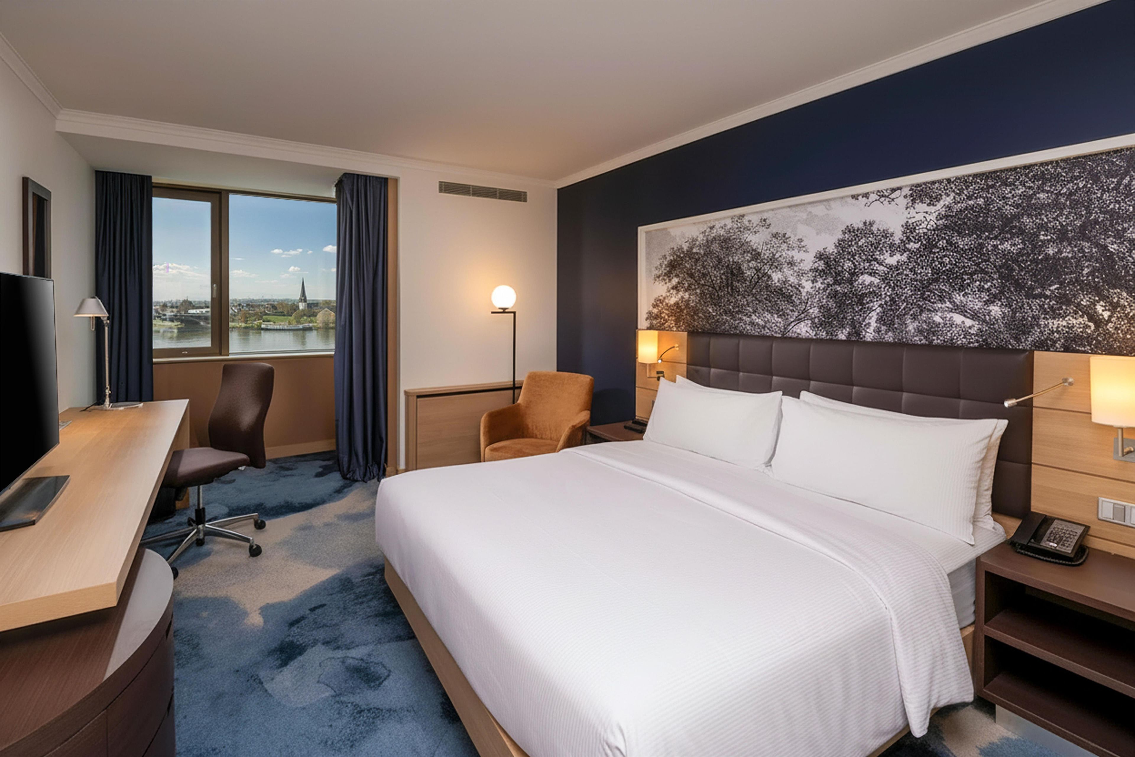 Habitación de hotel con una cama grande, un escritorio, una silla, una mesita de noche y vistas de un paisaje urbano a través de la ventana.