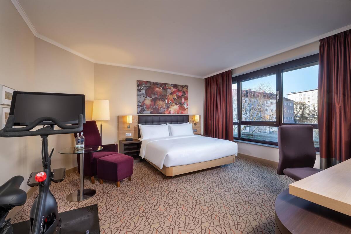 A hotel room with a bed, a desk, a chair, a TV, and a treadmill.
