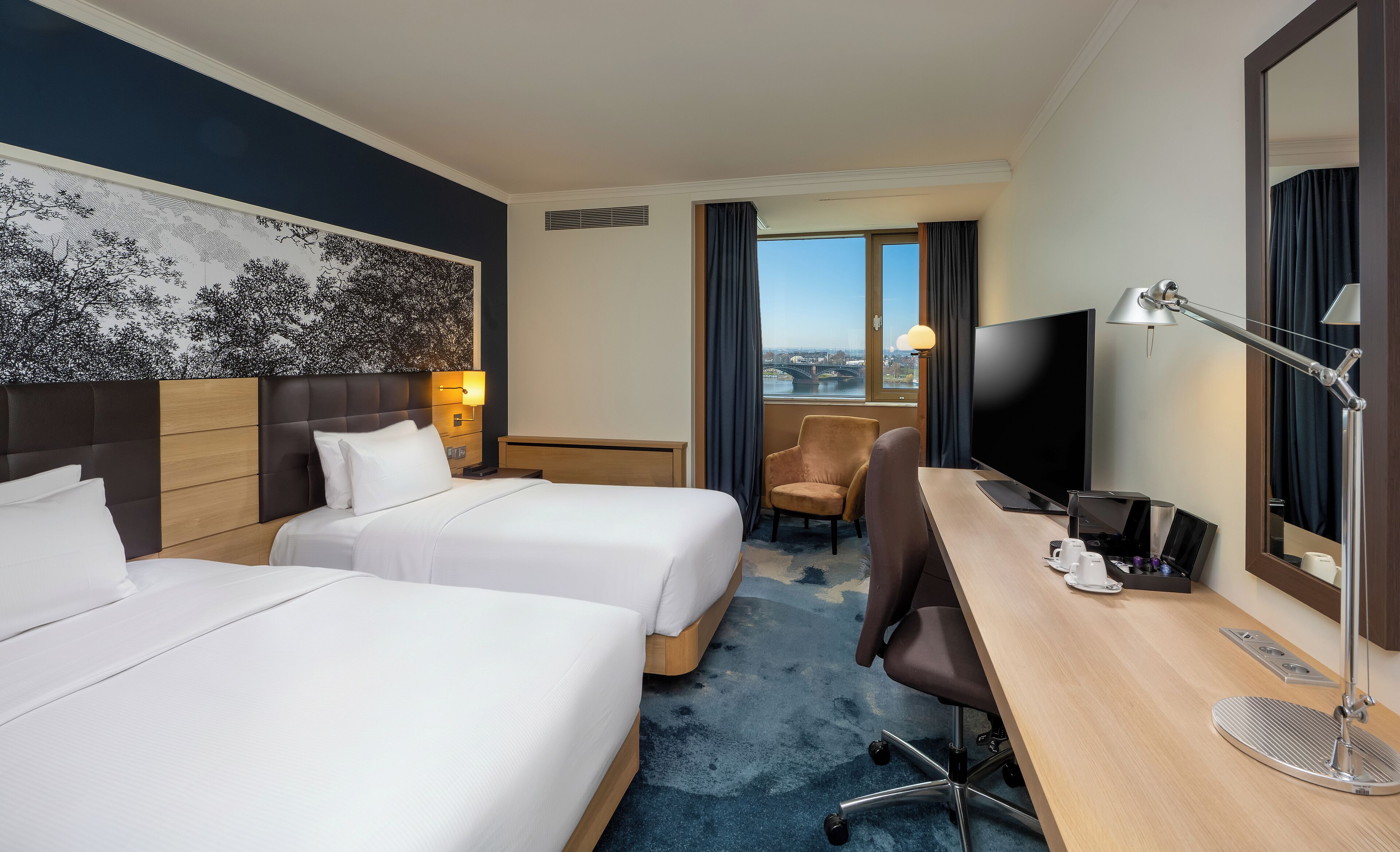 Habitación de hotel con dos camas, un escritorio con computadora, una silla, un televisor y un ventanal con vista a la ciudad.