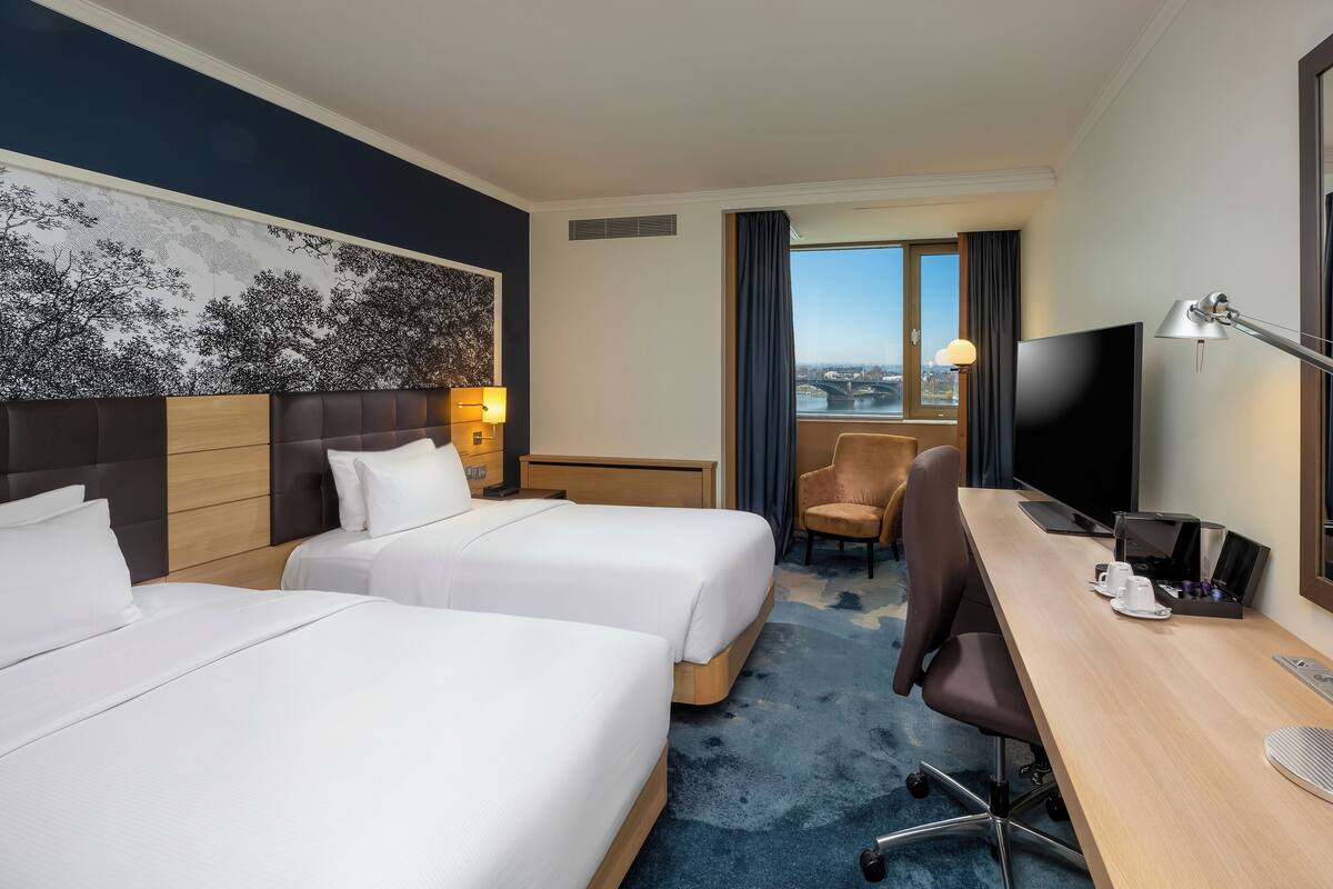 Habitación de hotel con dos camas, un escritorio con computadora, una silla, un televisor y un ventanal con vista a la ciudad.