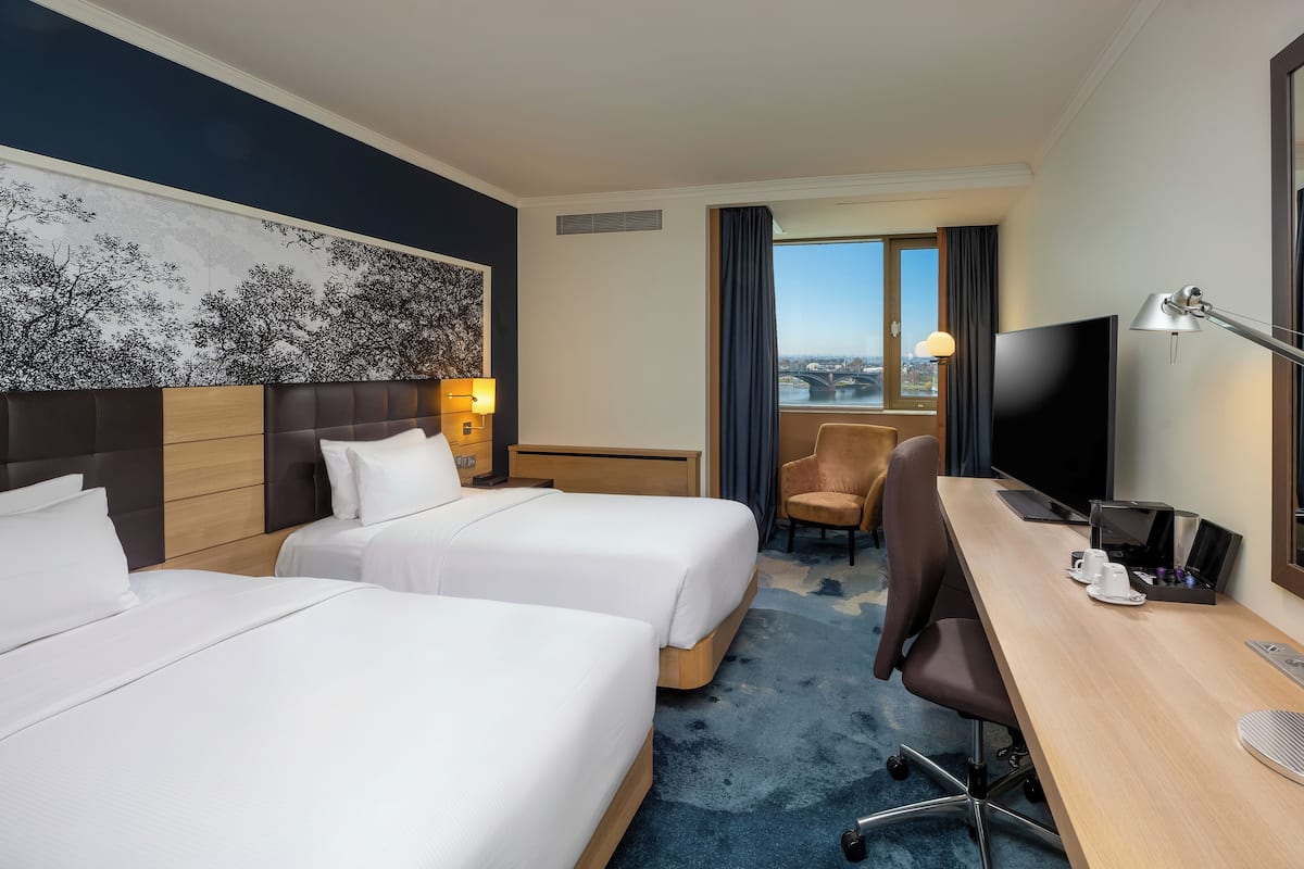 Habitación de hotel con dos camas, un escritorio con computadora, una silla, un televisor y un ventanal con vista a la ciudad.