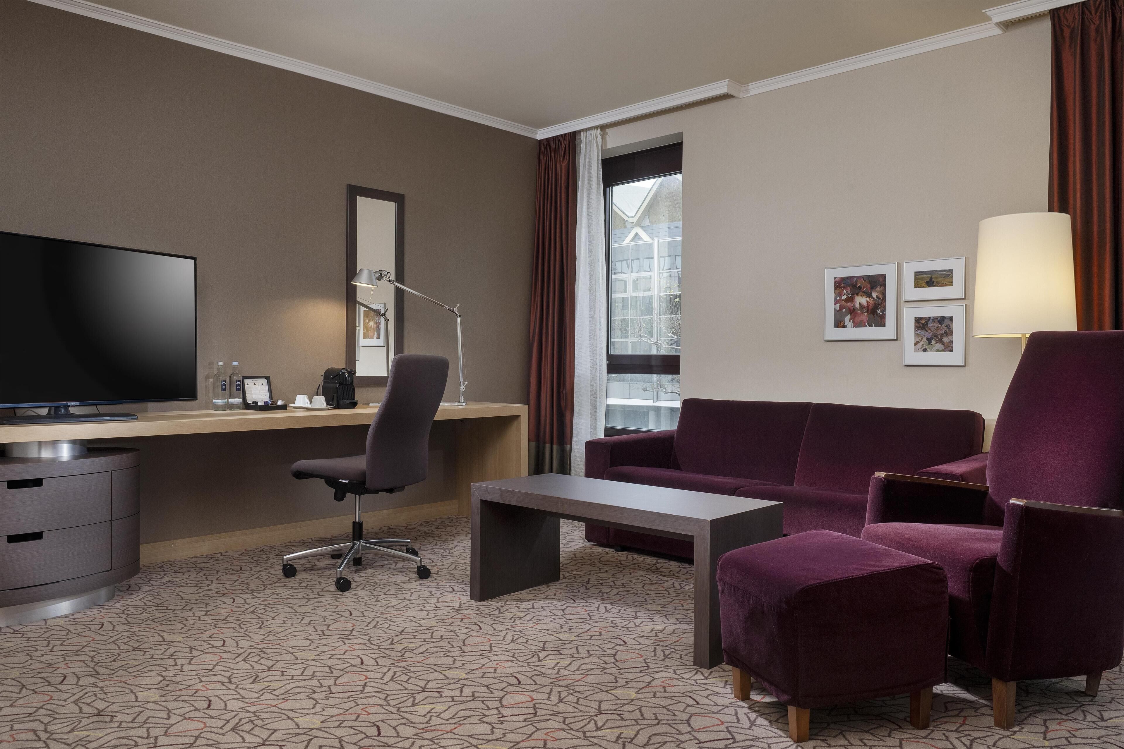 Ein Hotelzimmer mit Schreibtisch, Fernseher, Sessel, Schlafsofa, Lampe und einem Fenster mit Vorhängen.