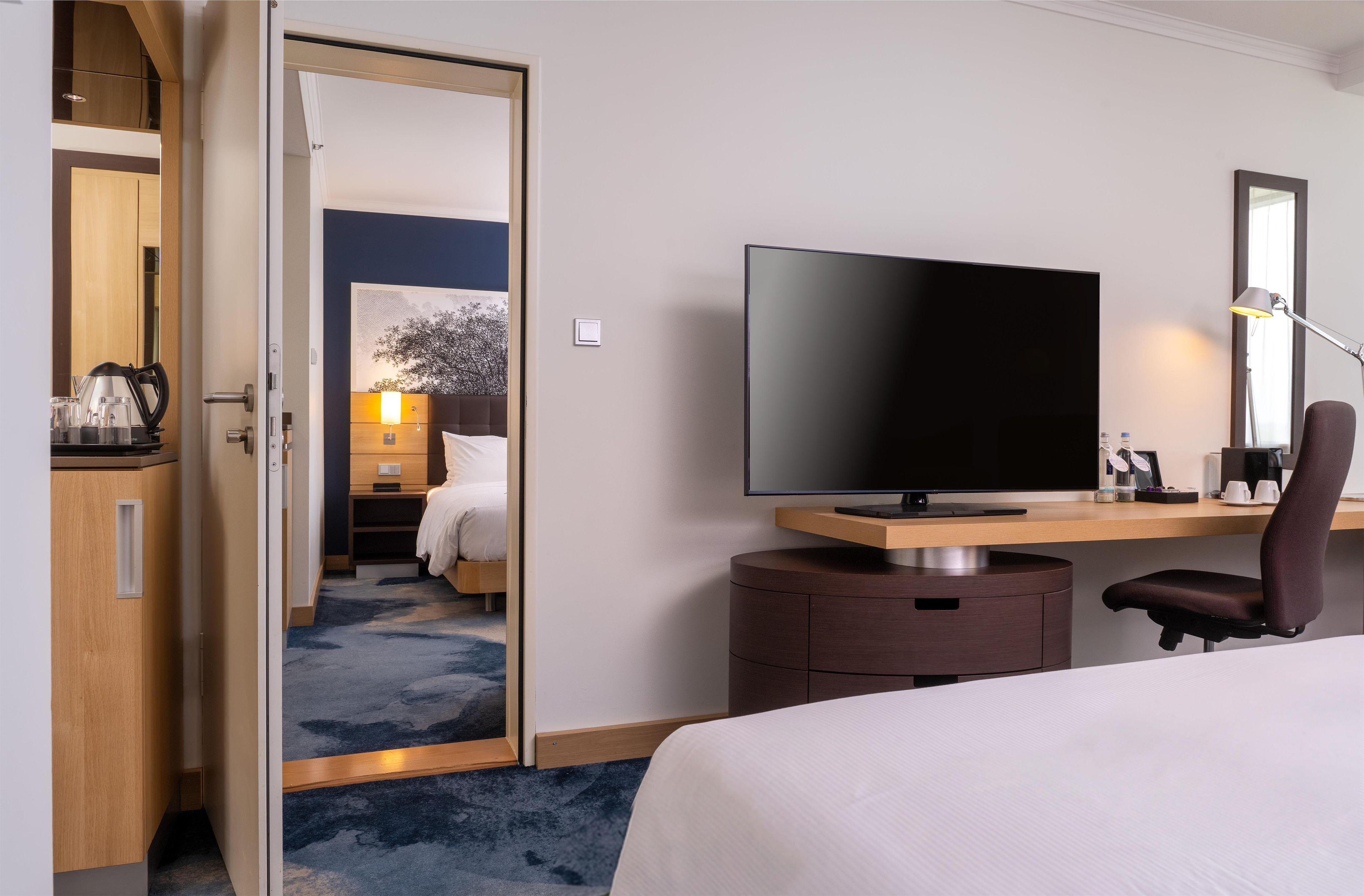 A hotel room with a bed, a desk with a flat-screen TV, a chair, a desk lamp, a mirror, and a wardrobe.