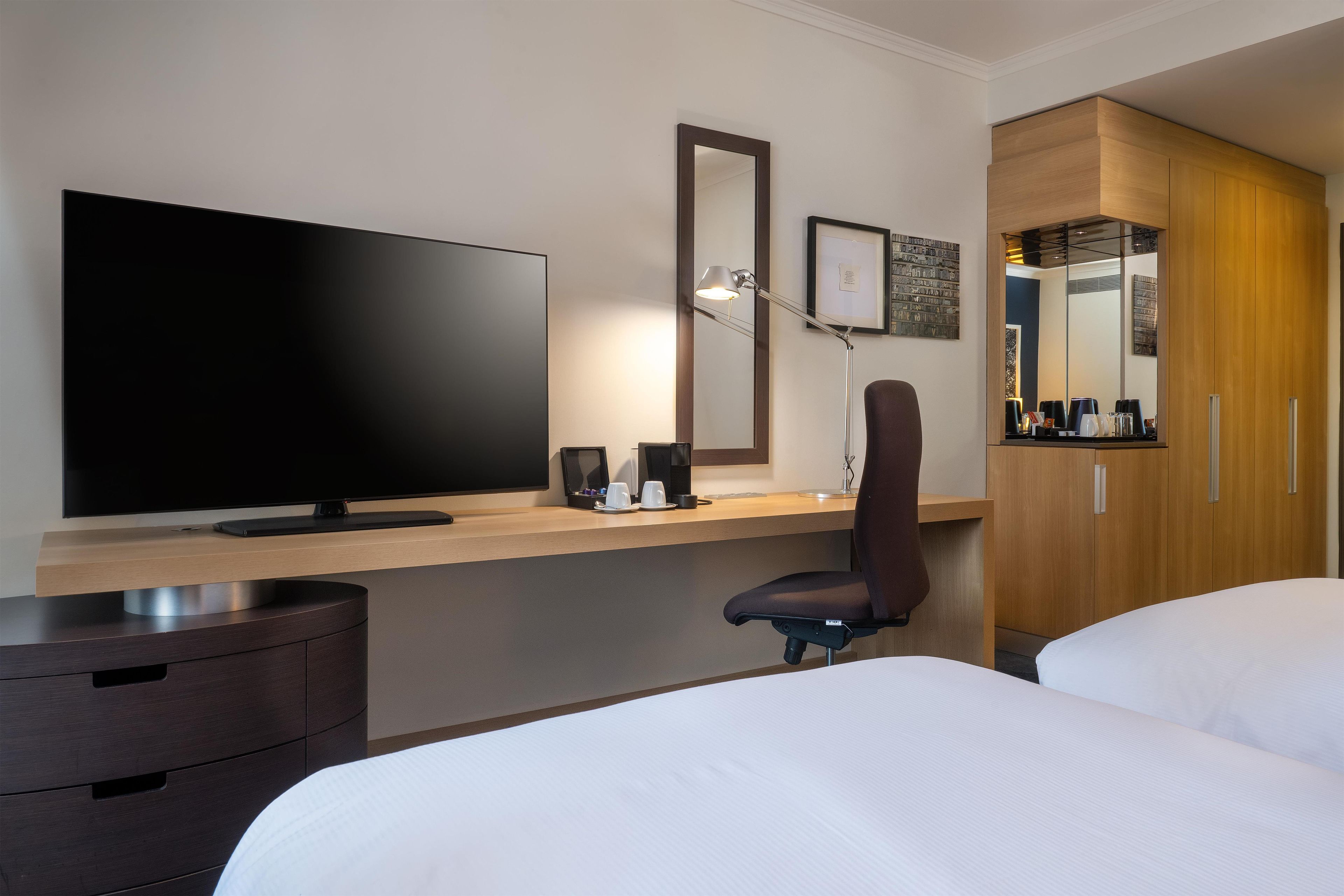 A hotel room with a flat-screen TV, a desk with a lamp, a chair, and a bed.