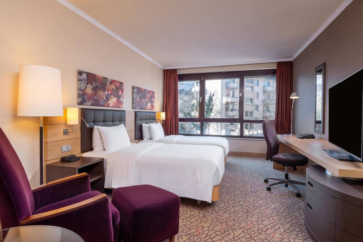 Habitación de hotel con dos camas, un escritorio, una silla, un televisor y obras de arte en las paredes.