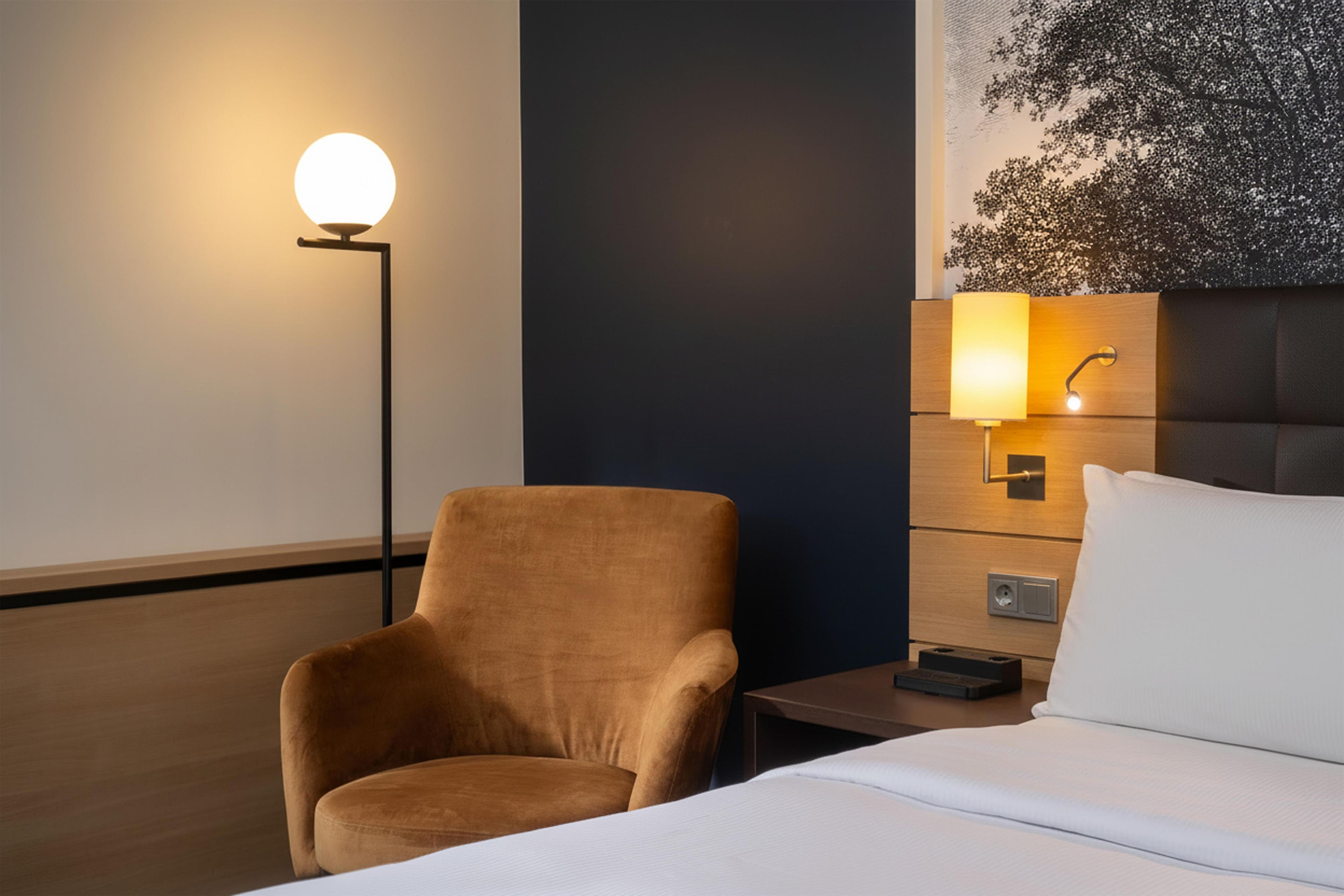 Une chambre d’hôtel avec un lit, une chaise, une table de chevet et une lampe fixée au mur.