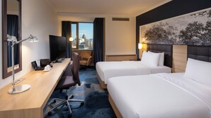 A hotel room with two beds, a desk with a computer, a TV, and a city view. - Hilton Mainz (Mainz)