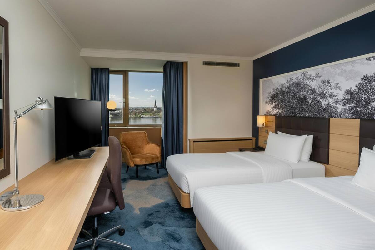 Habitación de hotel con escritorio, dos camas, una silla, televisor y vista a la ciudad.