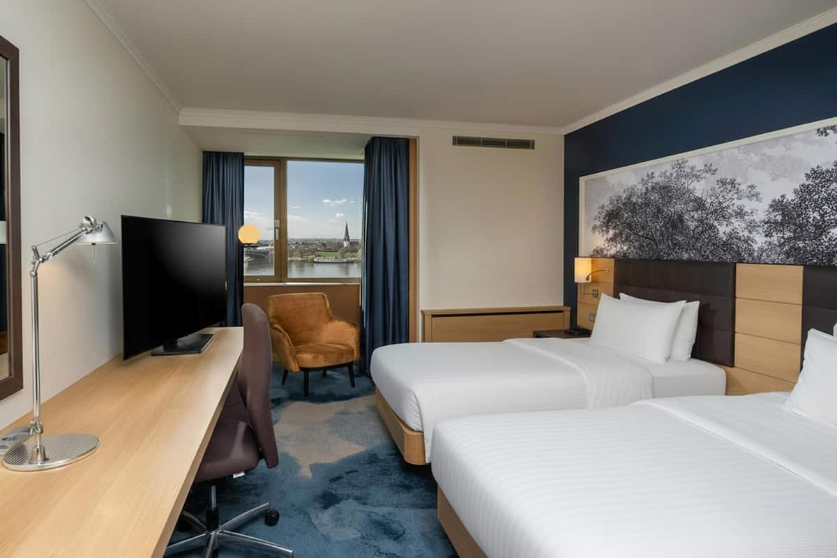 Habitación de hotel con escritorio, dos camas, una silla, televisor y vista a la ciudad.