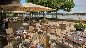 An outdoor dining area with tables and chairs, a bar, and a view of a river and bridge. - Hilton Mainz (Mainz)