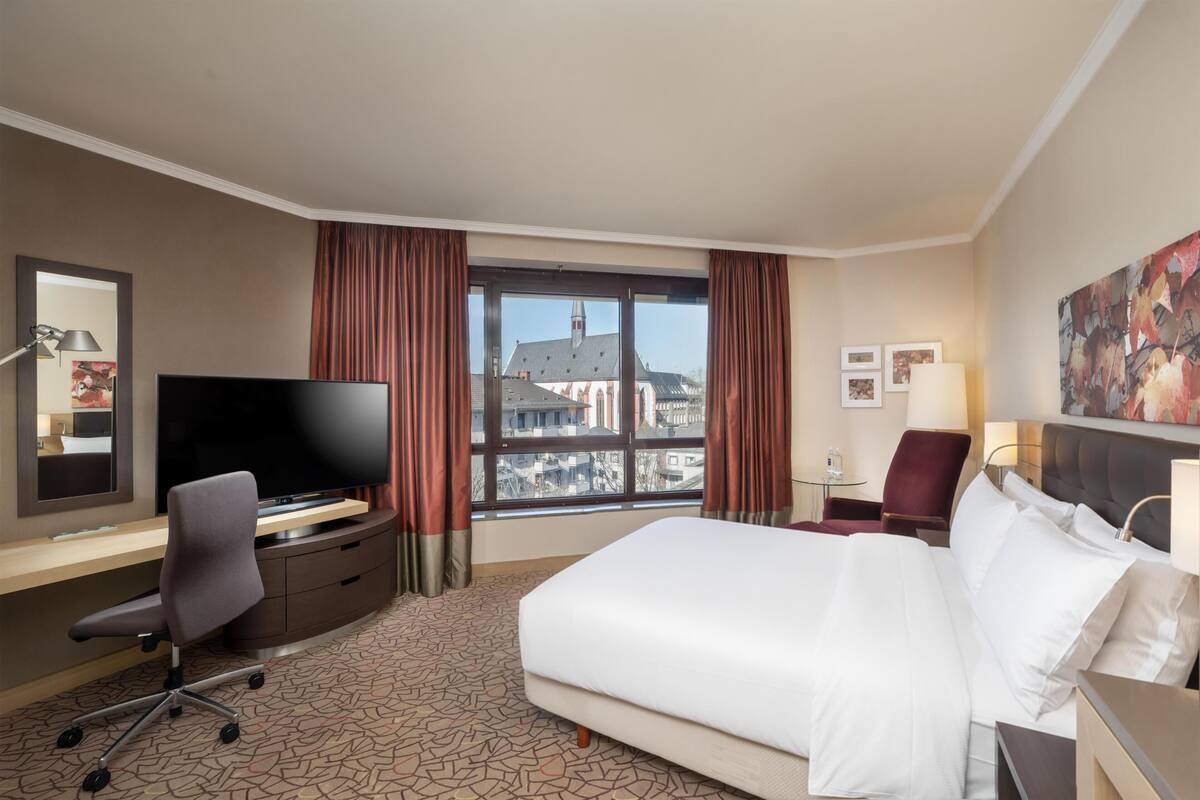 Habitación de hotel con cama, escritorio con televisor, una silla, ventana con cortinas y un cuadro en la pared.