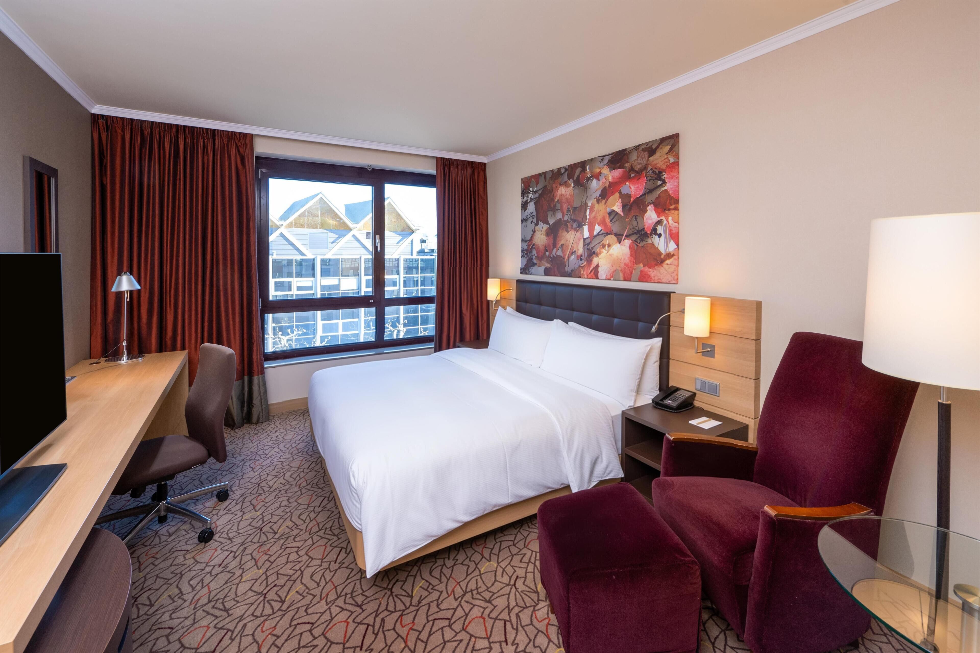 A hotel room with a bed, desk, chair, nightstand, lamp, and a window with curtains.