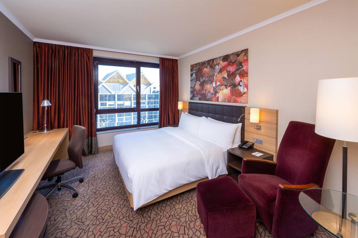 Una habitación de hotel con cama, escritorio, silla, mesita de noche, lámpara y ventana con cortinas.