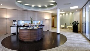 A modern reception area with a central counter, a coffee machine, a refrigerator, and a potted plant. - Hilton Mainz (Mainz)