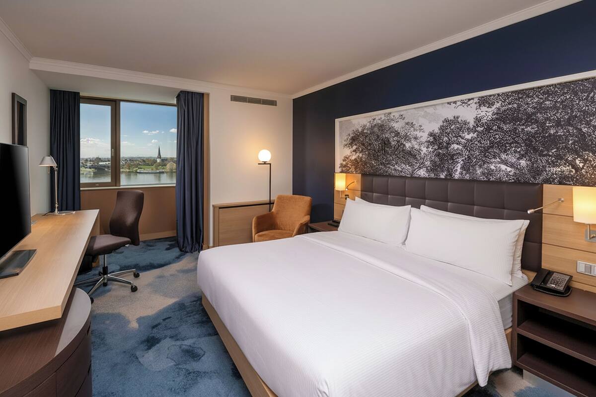 Habitación de hotel con una cama grande, un escritorio, una silla, un televisor y vistas de la ciudad a través del ventanal.