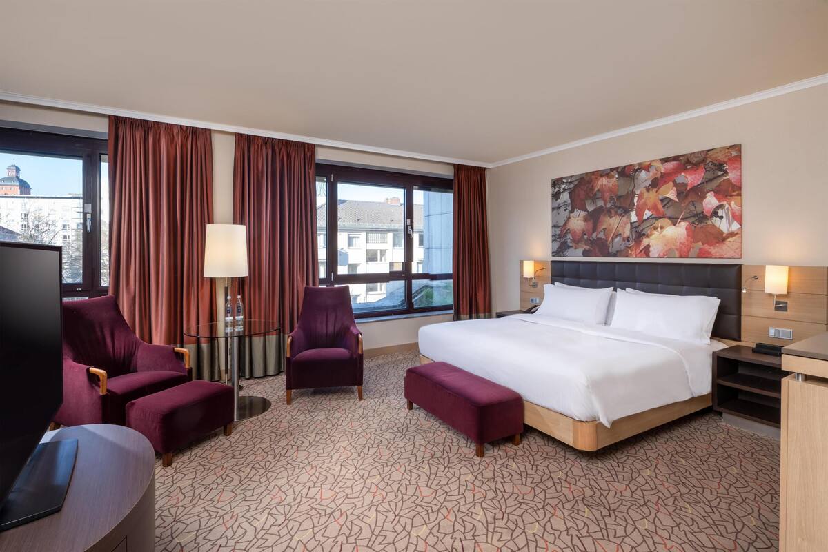 Habitación de hotel con una cama grande, dos sillones, un escritorio con una computadora portátil, un televisor y un amplio ventanal con cortinas.
