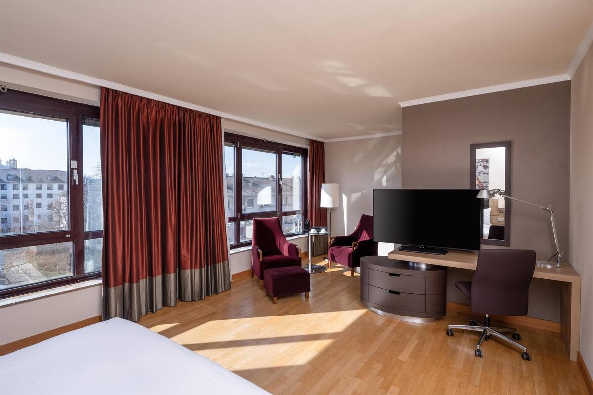 Una habitación de hotel con una cama, dos sillones, un escritorio con una computadora, un televisor y ventanas grandes con cortinas.