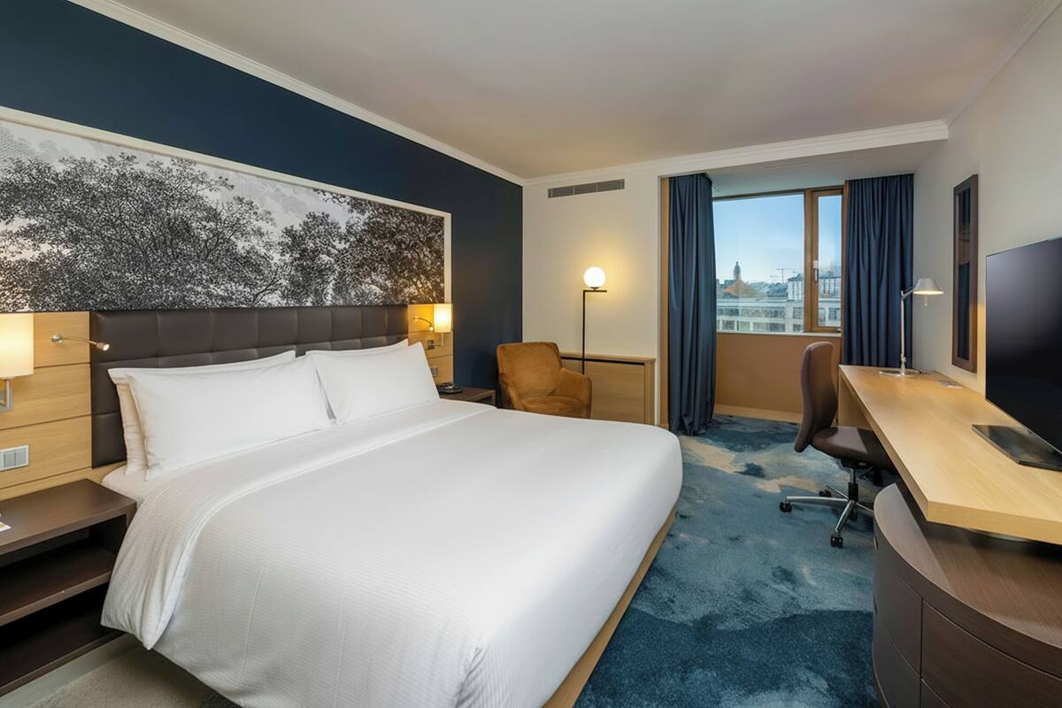 Habitación de hotel con una cama grande, un escritorio, una silla, un televisor y vistas a la ciudad.