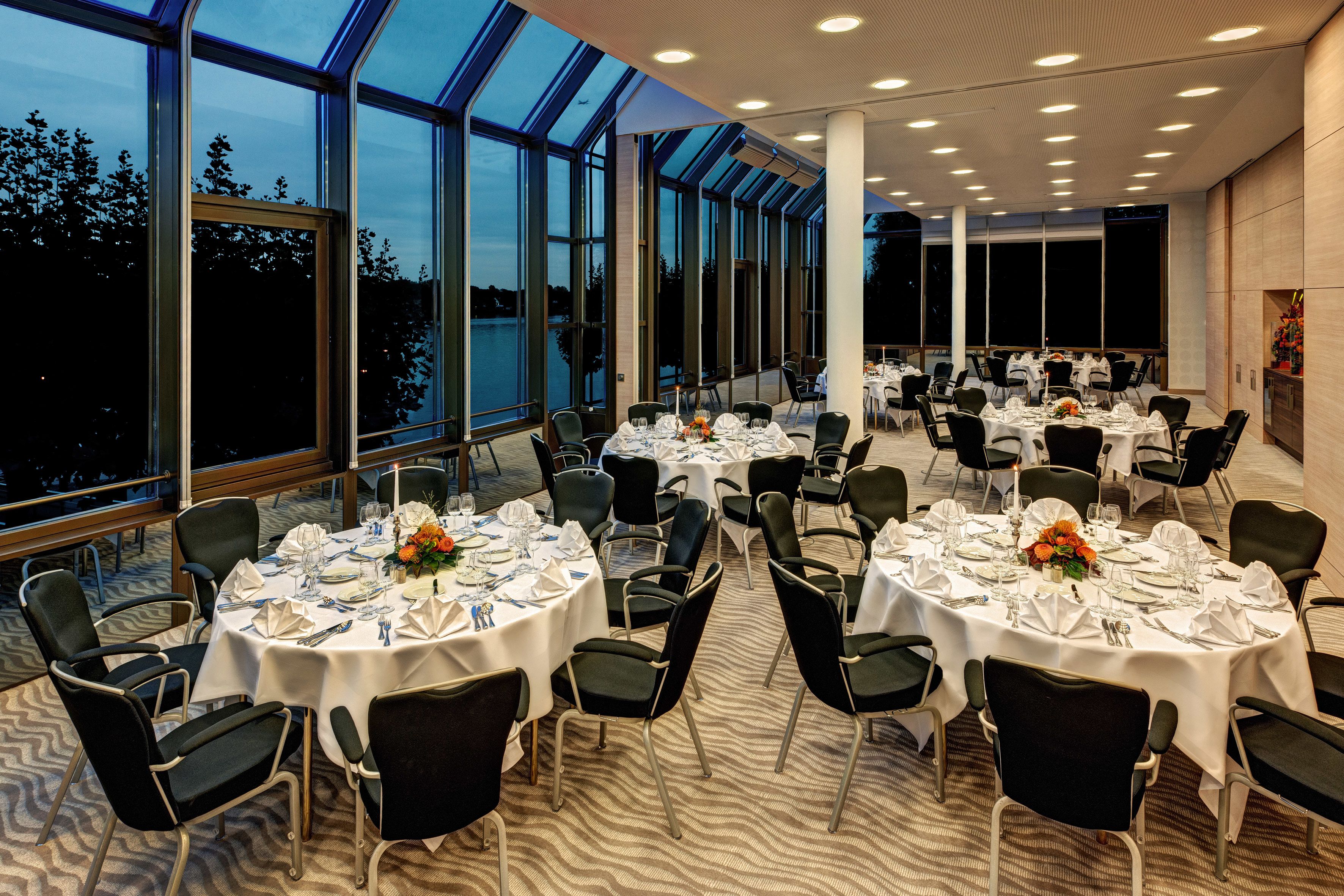 A spacious dining area with multiple round tables set for dining, large windows offering views of the outdoors, and a modern ceiling design.