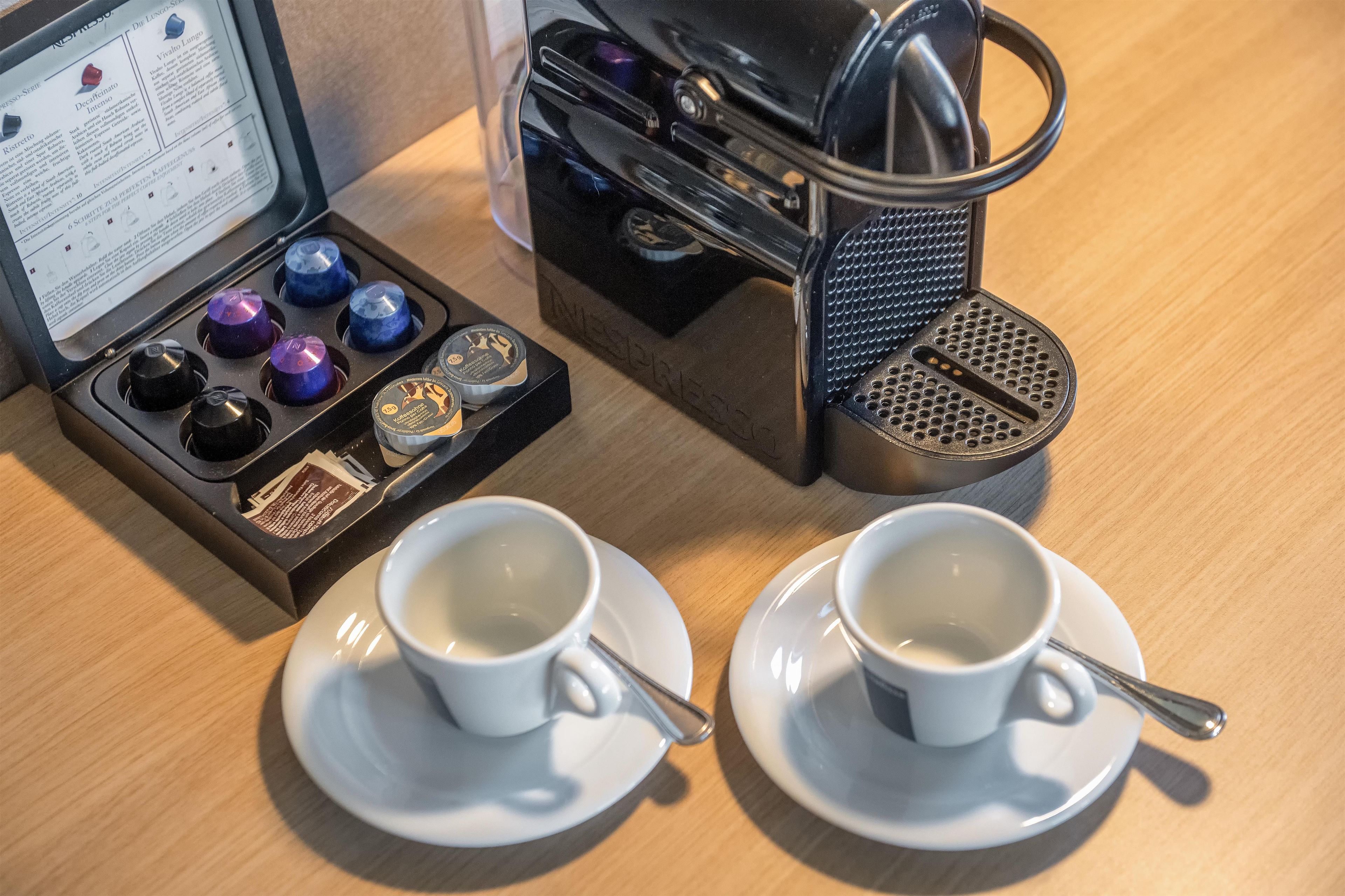 Una cafetera con su manual y cápsulas de café, dos tazas en platillos, y una cuchara.