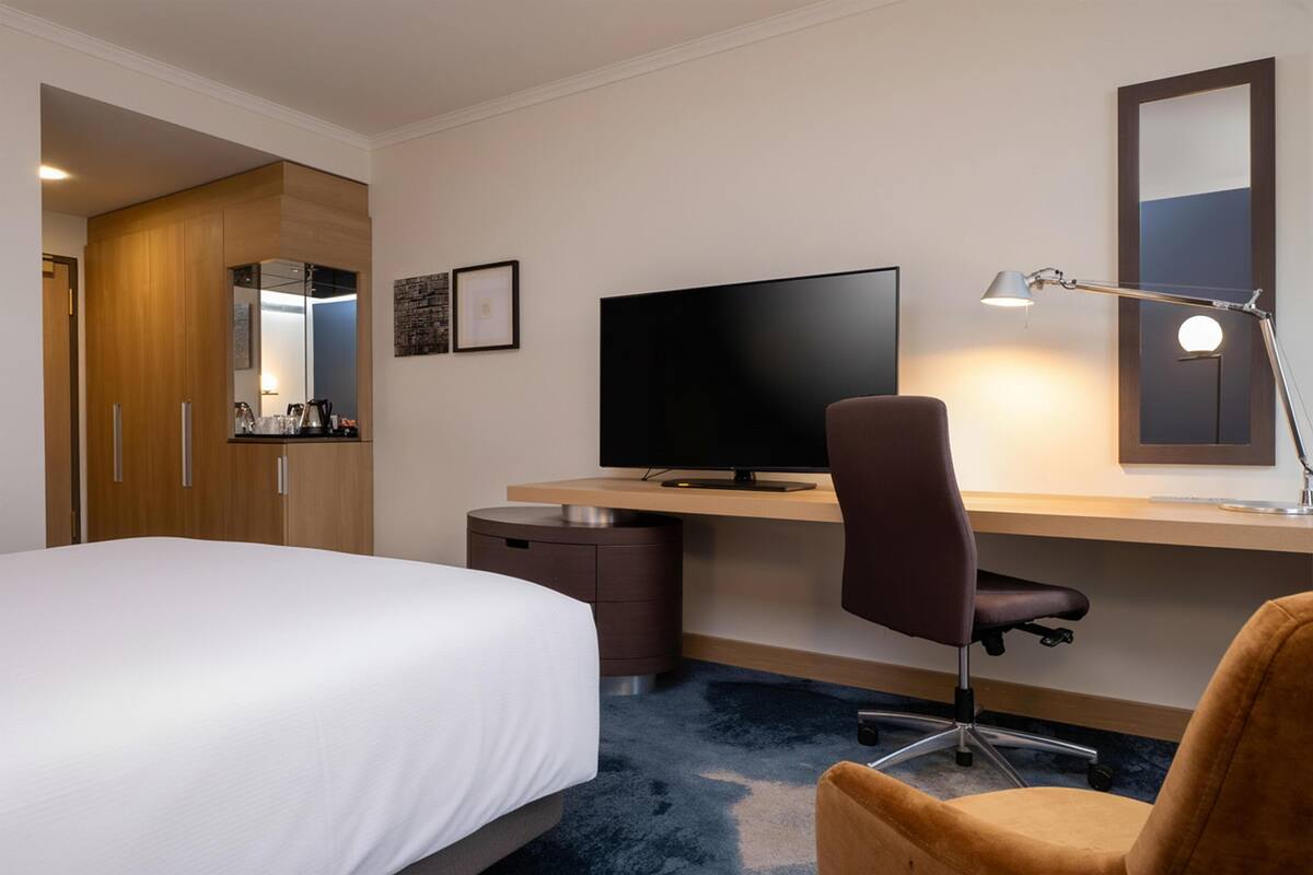 Una habitación de hotel con una cama, un escritorio con computadora, una silla, una lámpara de escritorio, un espejo y un armario.