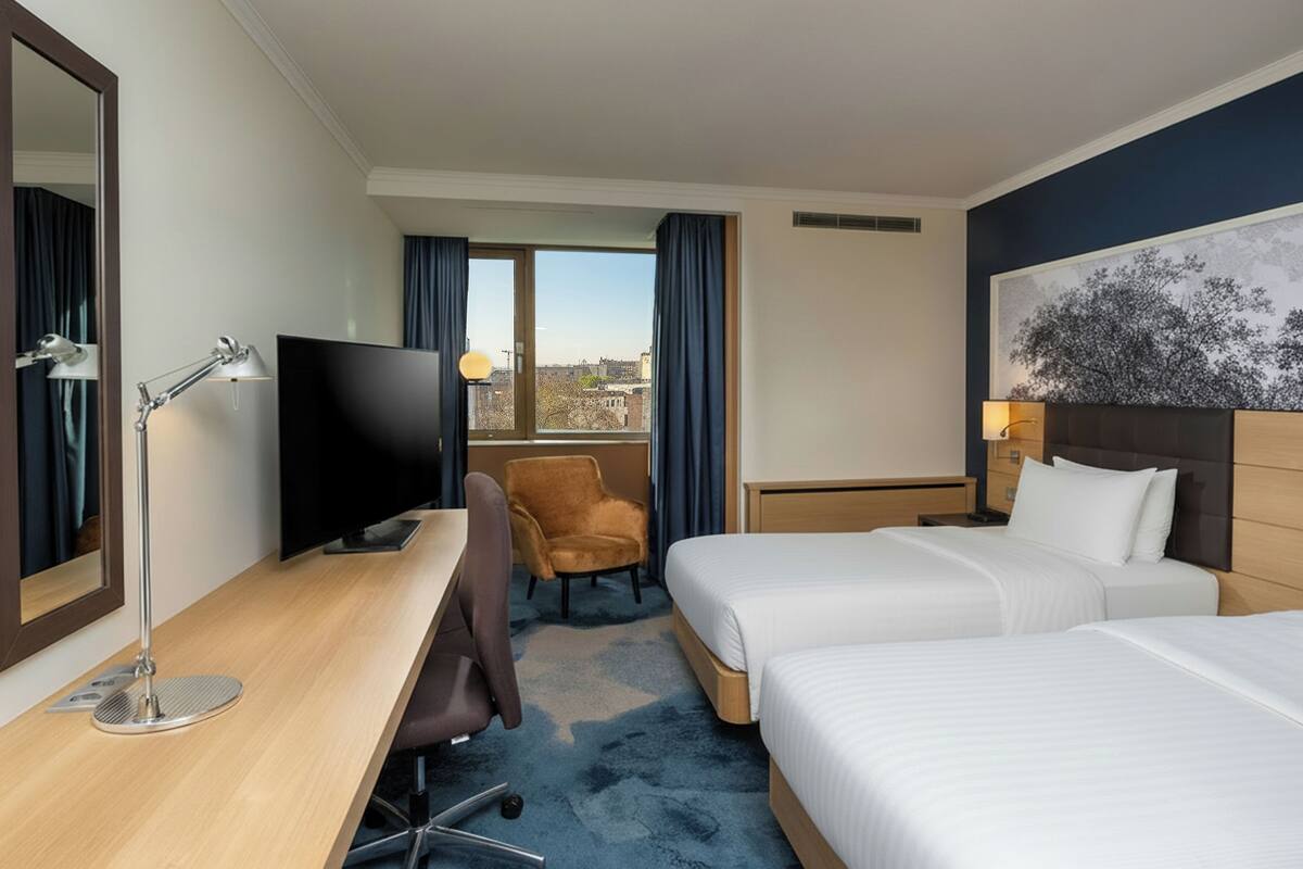Habitación de hotel con escritorio, dos camas, televisor, silla y ventana con cortinas.