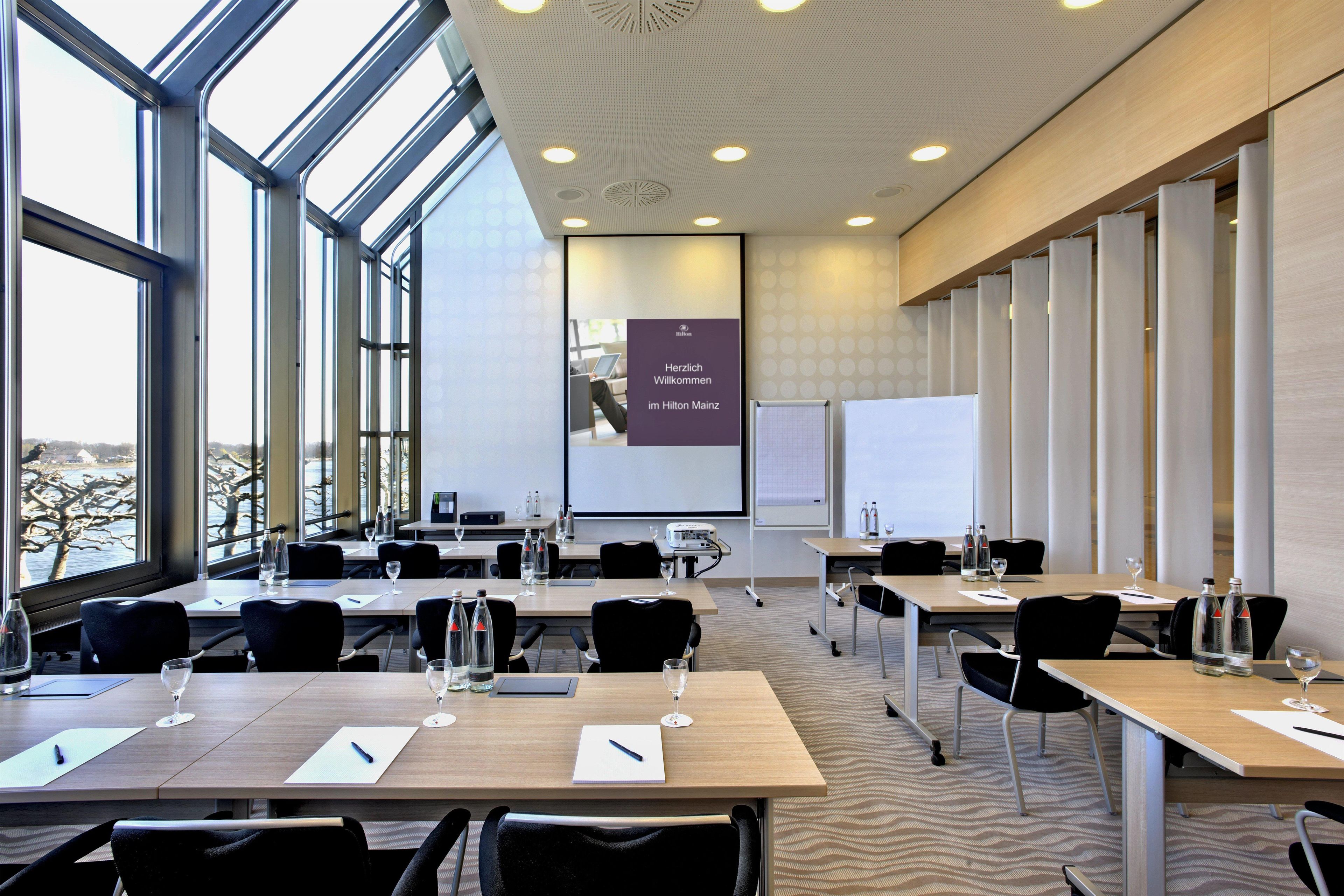 Un salón de conferencias con amplias ventanas, una pantalla de proyector y mesas equipadas con sillas y botellas de agua.