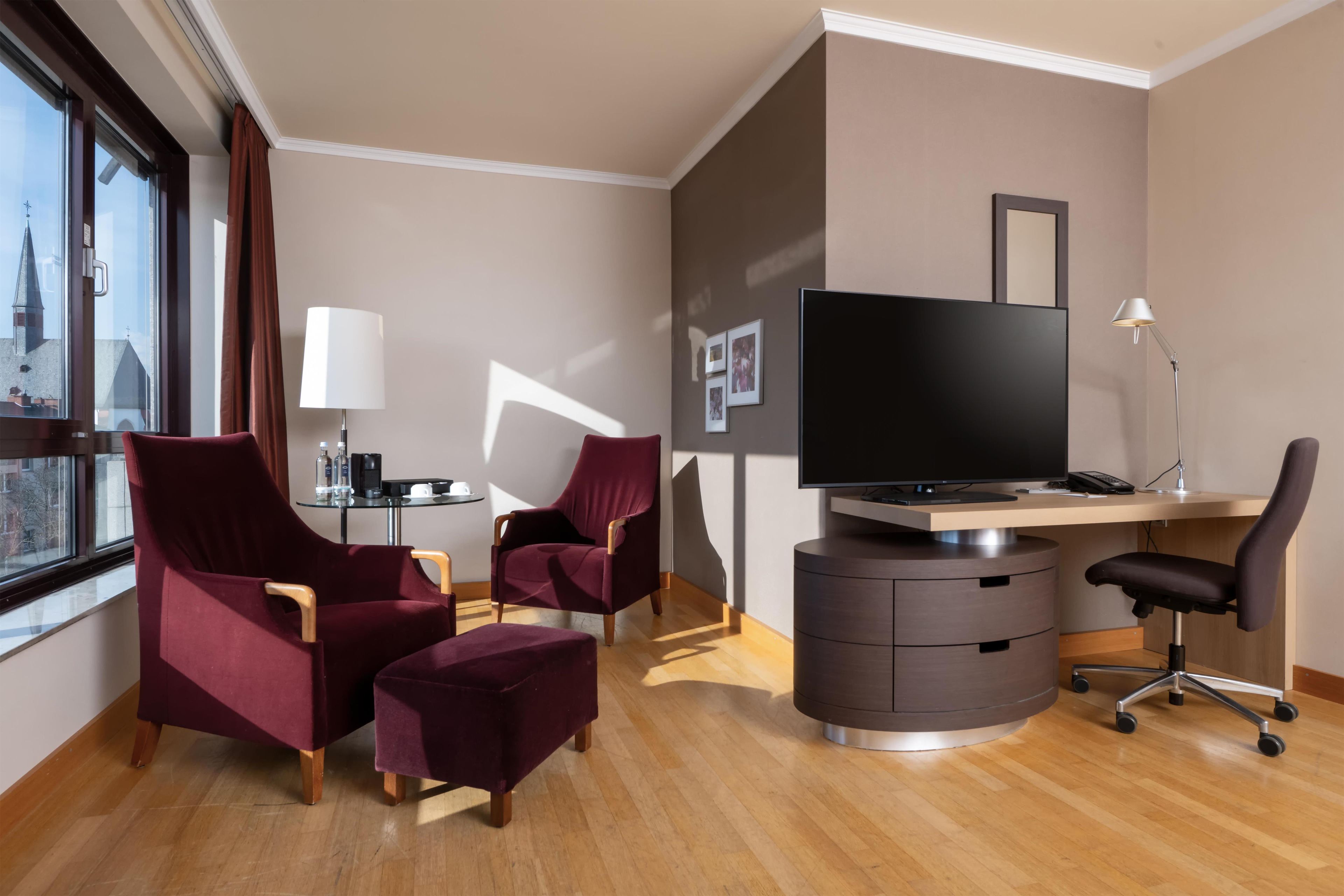 Une chambre d’hôtel avec une grande fenêtre, deux fauteuils rouges, une petite table ronde avec une lampe, un bureau avec un téléviseur à écran plat et une chaise.