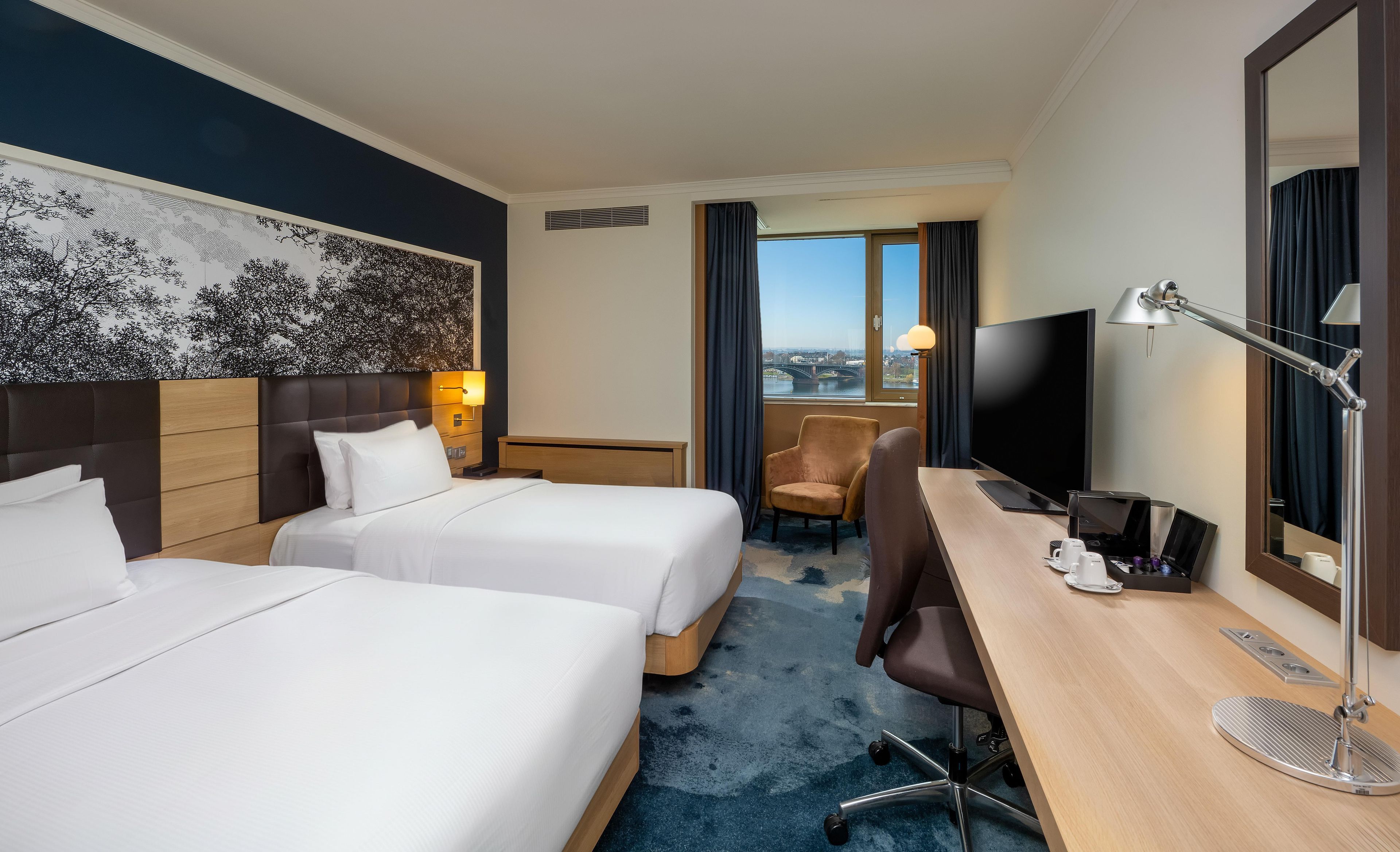 Ein Hotelzimmer mit zwei Betten, einem Schreibtisch, einem Sessel, einem Fernseher und einem großen Fenster mit Blick.