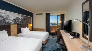 Ein Hotelzimmer mit zwei Betten, einem Schreibtisch, einem Sessel, einem Fernseher und einem großen Fenster mit Blick.