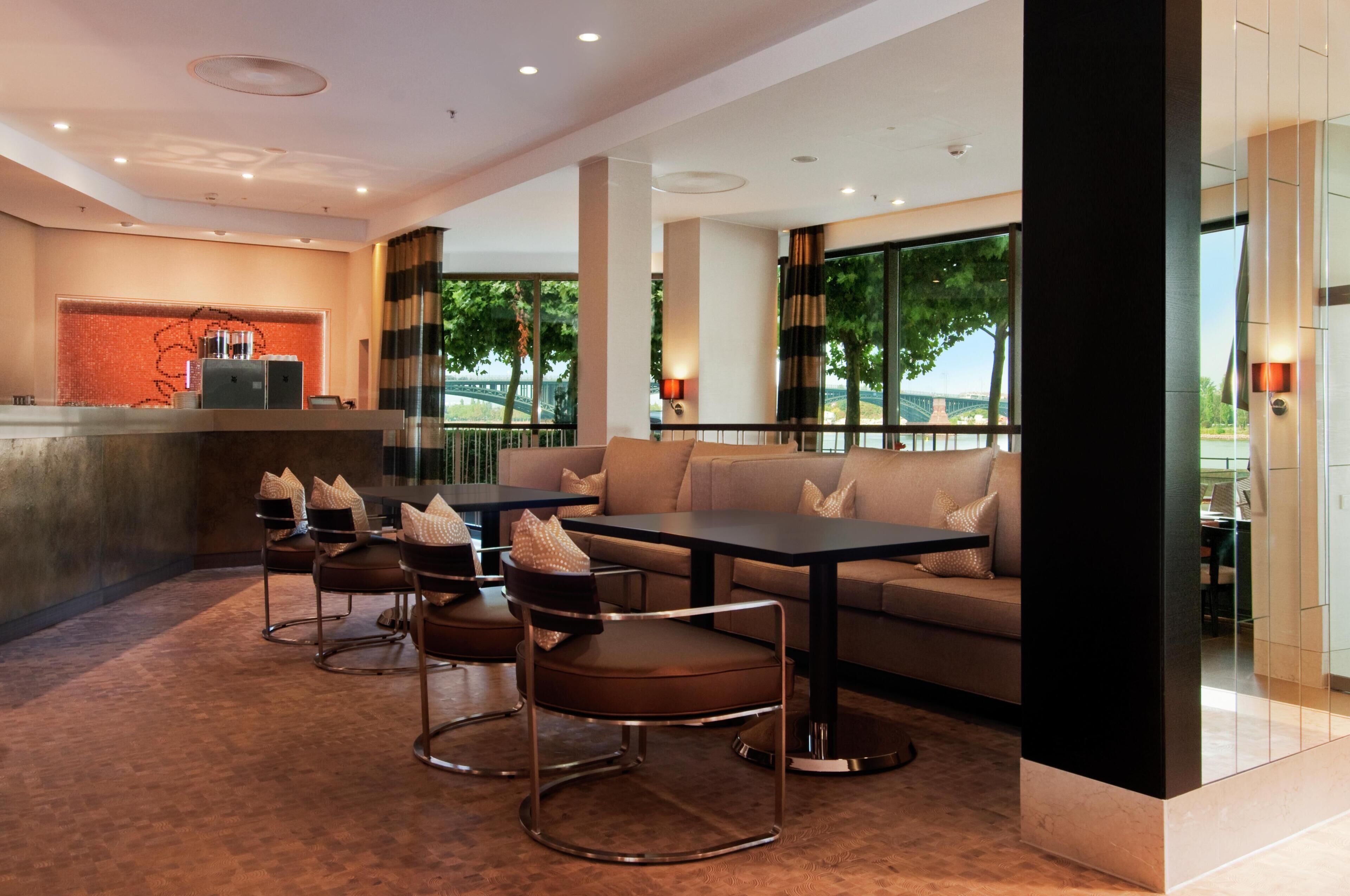 A modern hotel lobby with a reception desk, seating area, and large windows offering a view of greenery and a bridge.